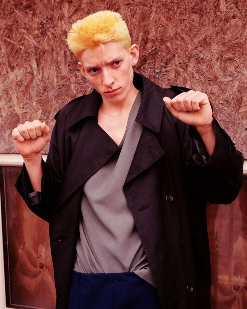 Bleached blonde male model posing with raised fists in a black trench coat and grey top against a wooden wall.