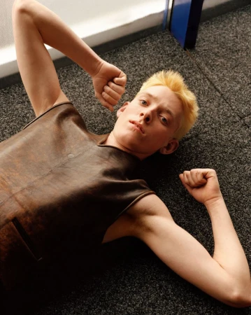 Blond model lying on a dark carpeted floor wearing a sleeveless brown leather top with arms raised.