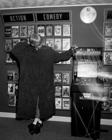Model in an oversized long dark coat posing beside an arcade cabinet in front of VHS tape shelves in black and white.