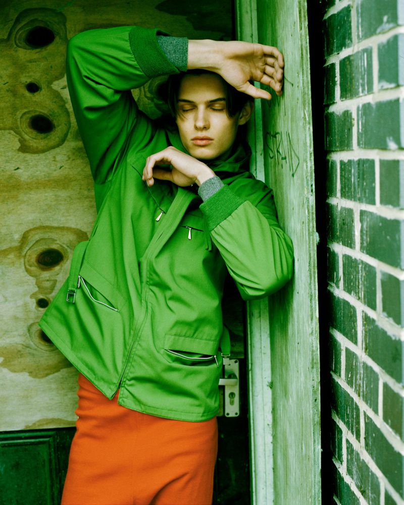 Young man wearing a bright green zip jacket and orange trousers leaning against a weathered green doorway beside a green brick wall.