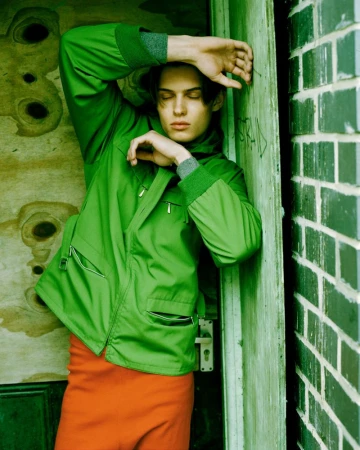 Young man wearing a bright green zip jacket and orange trousers leaning against a weathered green doorway beside a green brick wall.