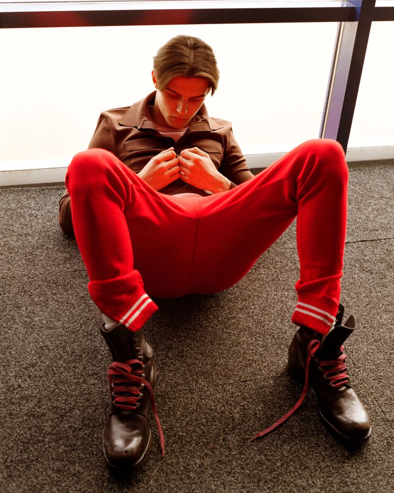 Young man in a brown shirt, red track pants, and brown lace-up boots sitting on a carpeted floor leaning against a window frame.
