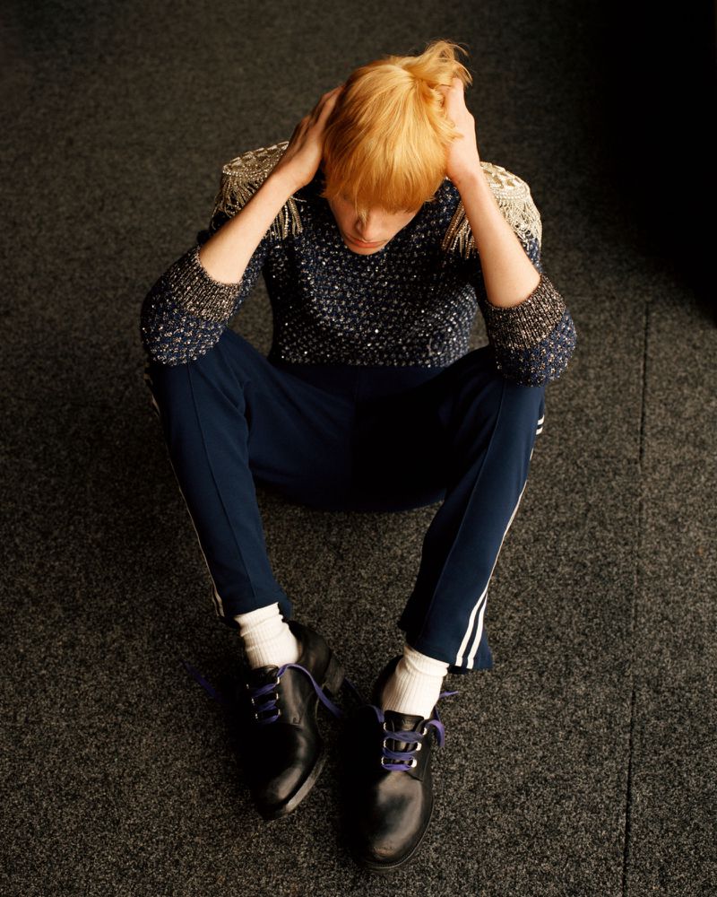High-angle view of a blonde model seated on a dark textured carpet wearing an embellished sweater, navy track pants, and black shoes.
