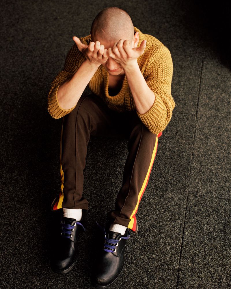 Man with a buzz cut seated on dark carpet wearing a mustard knit sweater and brown track pants with hands covering his eyes.