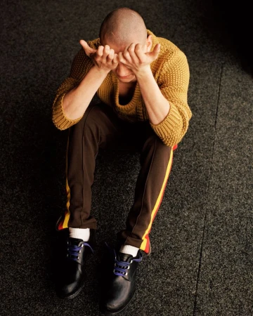 Man with a buzz cut seated on dark carpet wearing a mustard knit sweater and brown track pants with hands covering his eyes.