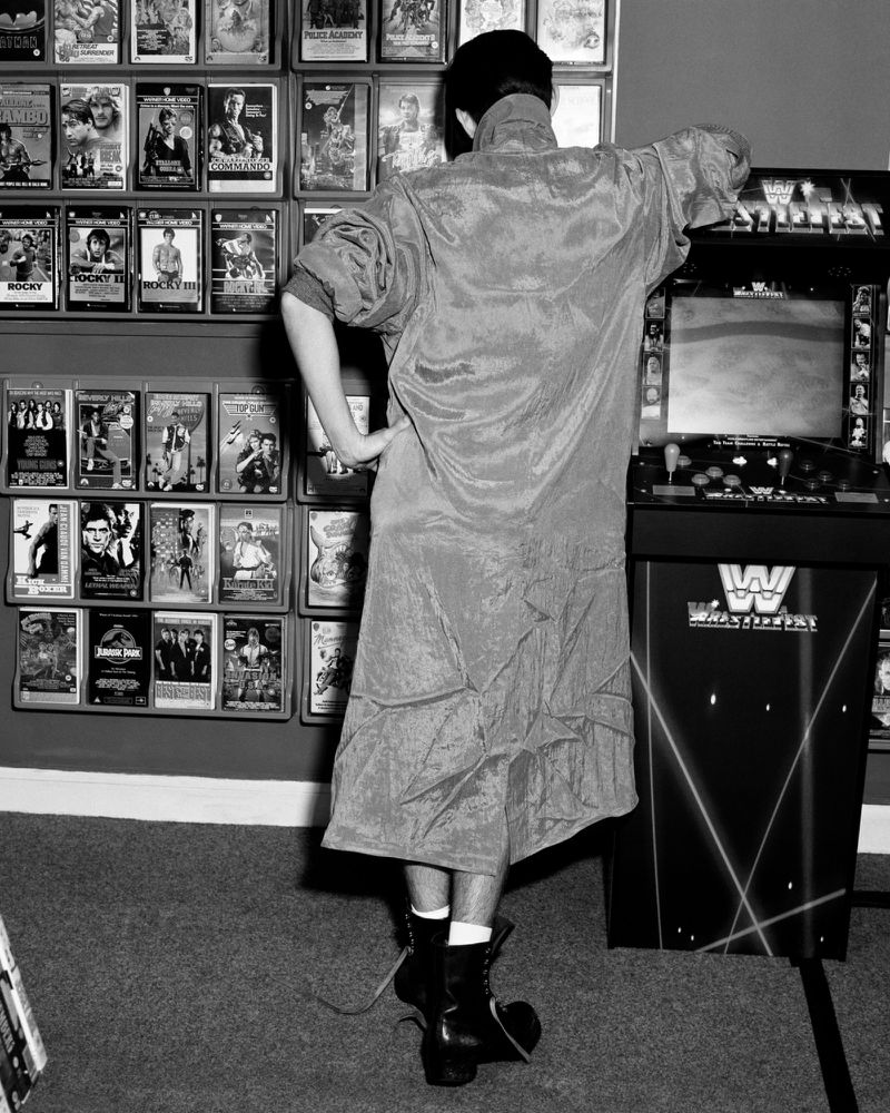 Monochrome image of a person wearing a long coat standing before arcade cabinets and video shelves for Arena Homme +