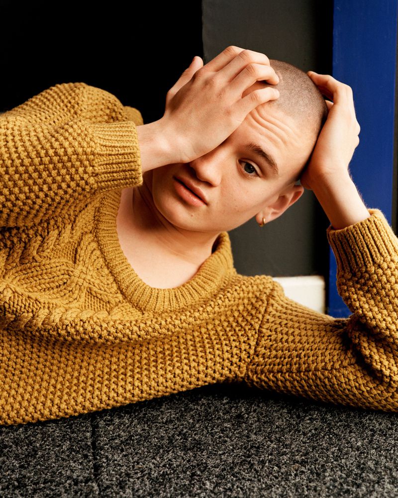 Model with a shaved head wearing a mustard yellow knit sweater reclines on a dark carpet with hand to forehead.