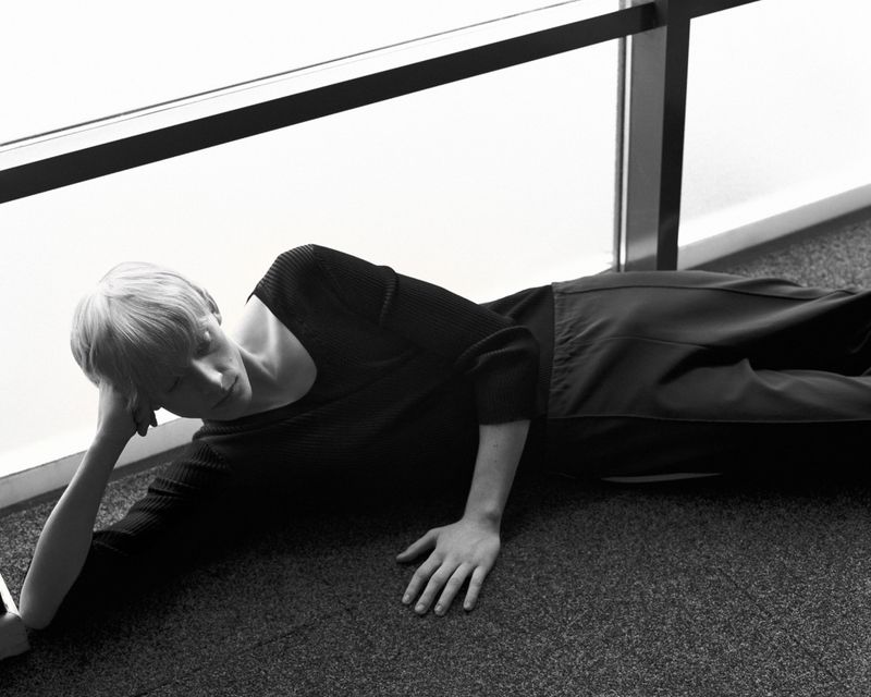 Model with short blonde hair lying on a carpeted floor wearing a black knit sweater and trousers next to a large window frame.