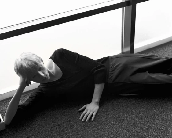 Model with short blonde hair lying on a carpeted floor wearing a black knit sweater and trousers next to a large window frame.
