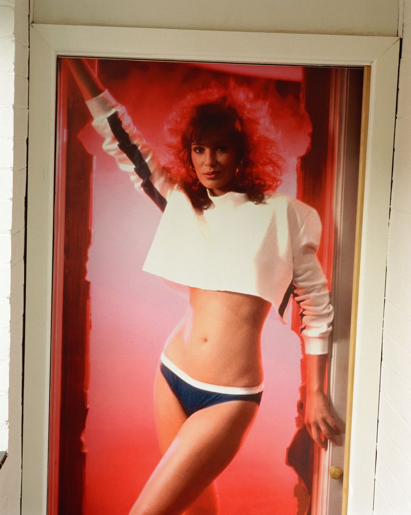 Model with voluminous auburn hair wearing a white cropped long-sleeve sweatshirt and navy briefs framed in a doorway against red light.