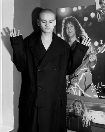 Monochrome portrait of a young man with a shaved head in an oversized black coat posing in front of music posters.