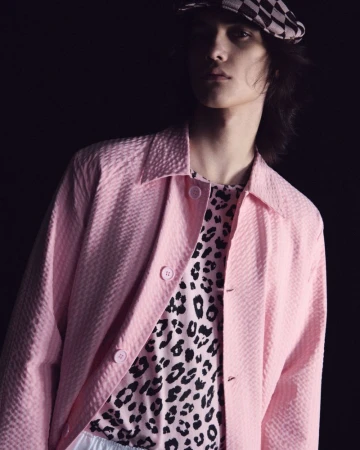 Model Nik Dermendzhiev in a light pink textured jacket, animal-print shirt, and checkered cap against a black studio background.