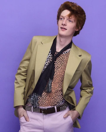 Redhead male model posing in a tan blazer, animal print top, and pink trousers against a purple backdrop