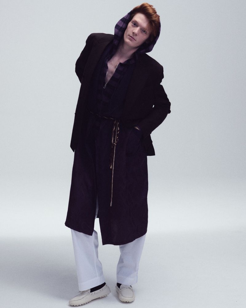 Male model with red hair posing in a long dark coat, hooded top, and white trousers against a white seamless studio background.