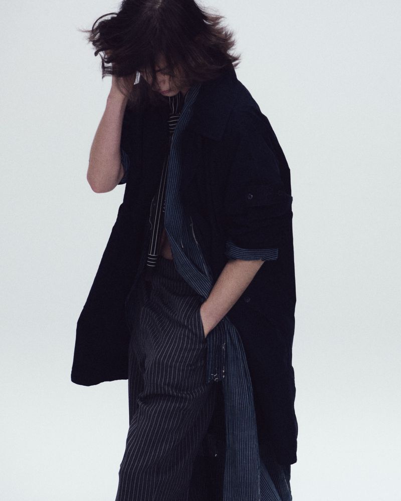 Model in a dark oversized coat and striped trousers looking downward against a seamless white studio background.