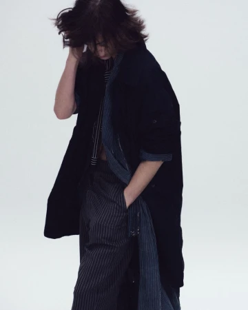 Model in a dark oversized coat and striped trousers looking downward against a seamless white studio background.