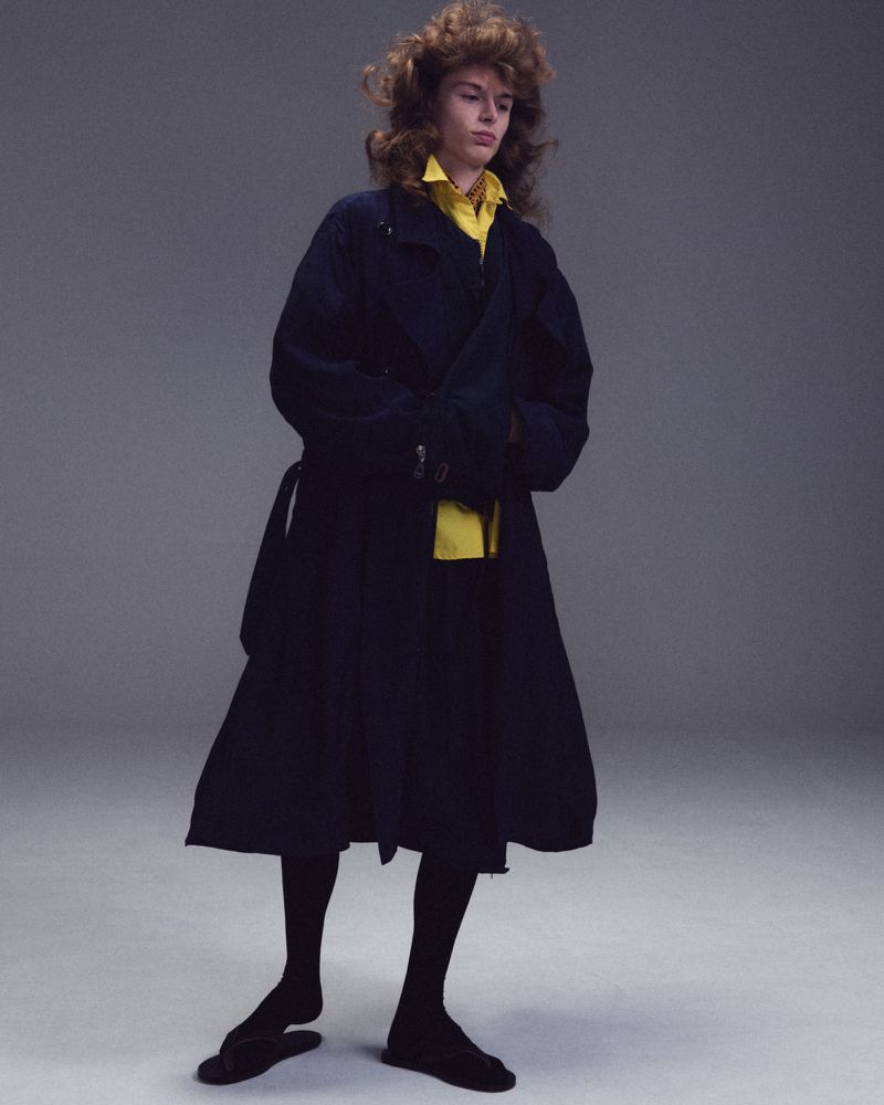 Model Nik Dermendzhiev wearing a long black coat over a yellow collared shirt with tights against a grey studio backdrop.