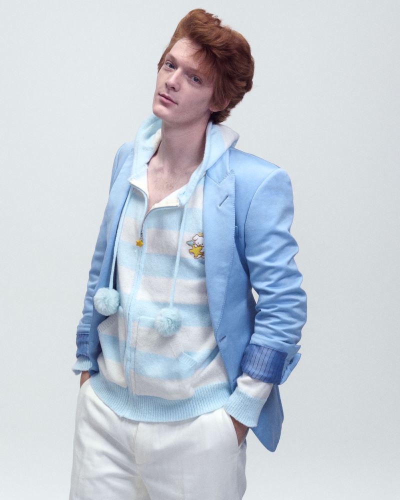 Model Nik Dermendzhiev wearing a light blue blazer over a striped pastel hoodie and white trousers against a white backdrop.