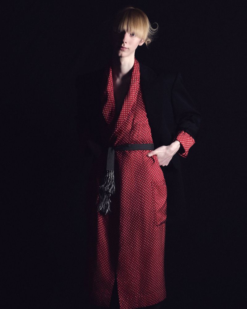 Nik Dermendzhiev wearing a red polka dot belted robe and black blazer against a dark studio background.