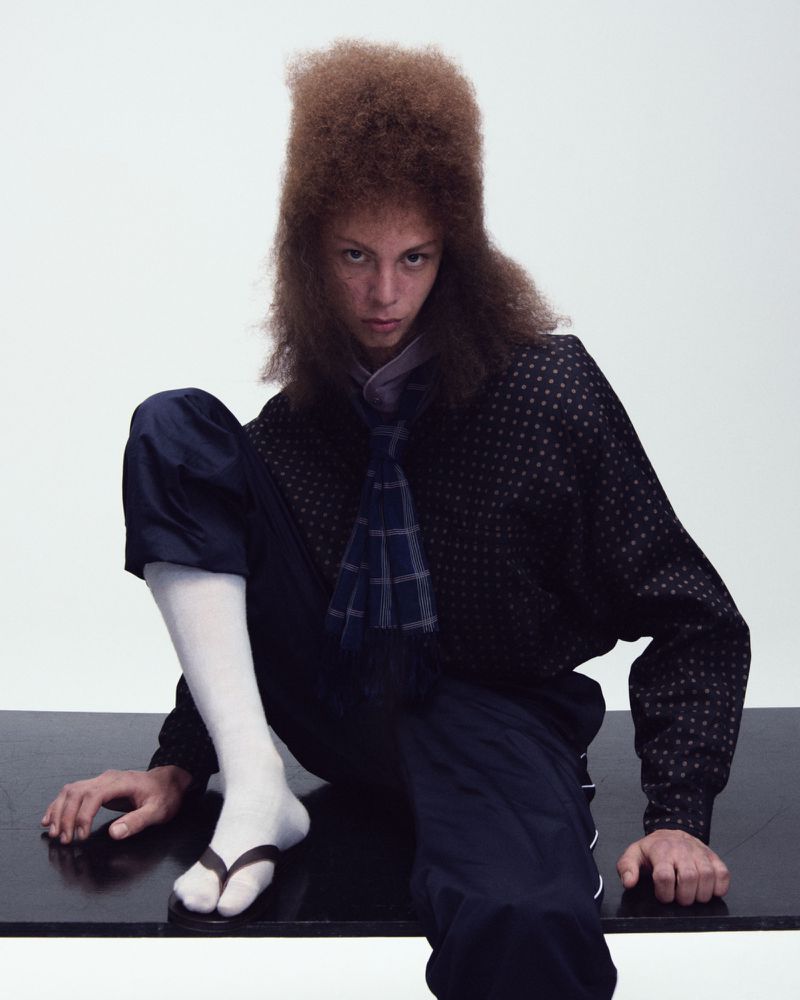 Model with tall brown hair wearing a patterned dark shirt and white socks with sandals against a white backdrop.