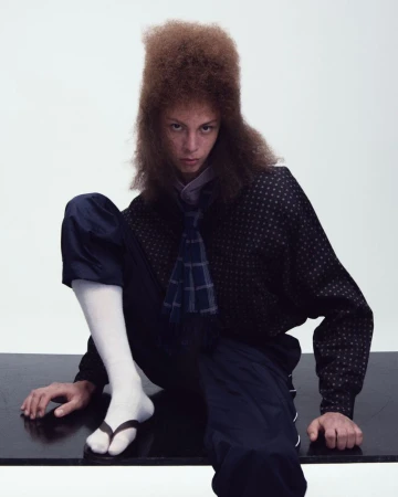 Model with tall brown hair wearing a patterned dark shirt and white socks with sandals against a white backdrop.