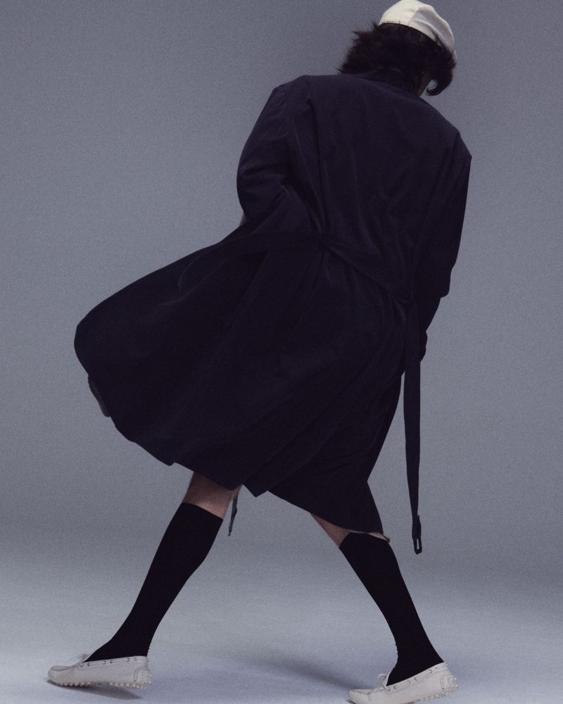 Model seen from behind wearing an oversized black coat, white cap, black socks, and white shoes against a grey studio backdrop.