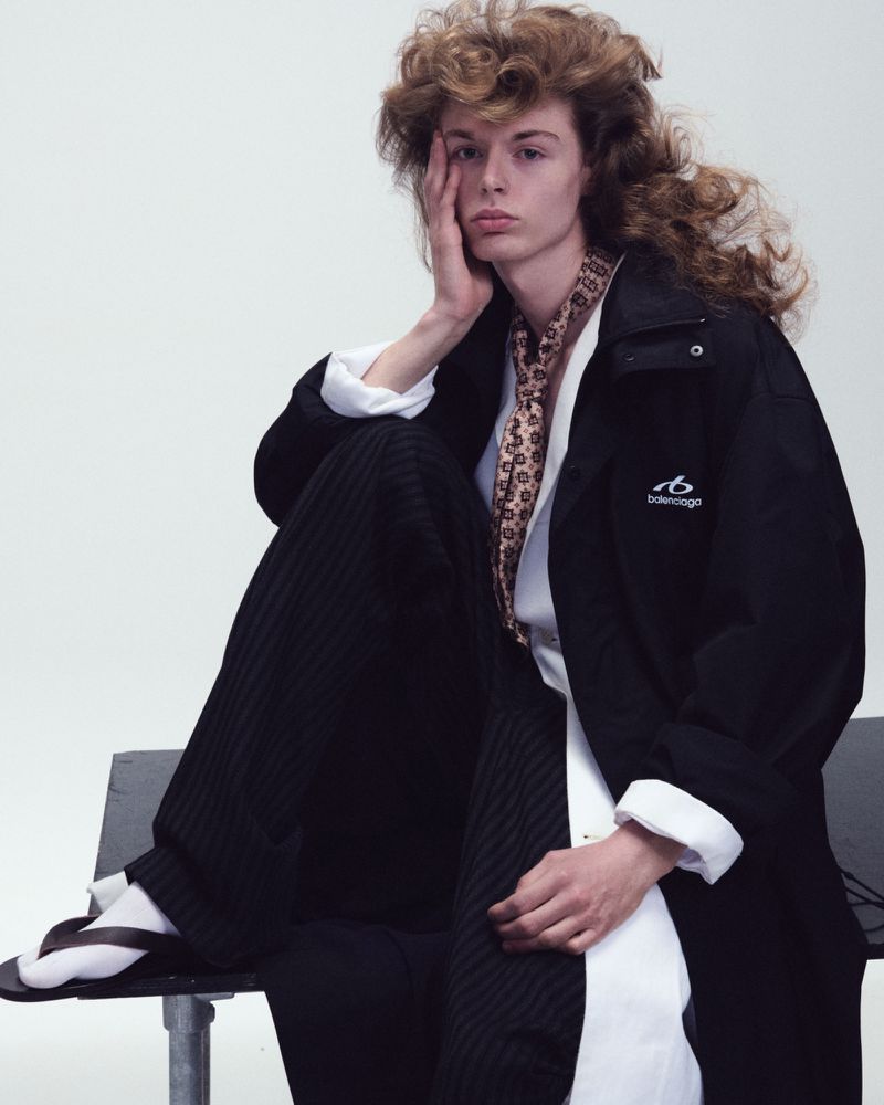 Model Nik Dermendzhiev wearing a black Balenciaga coat, patterned scarf, and white shirt seated in front of a white studio backdrop.