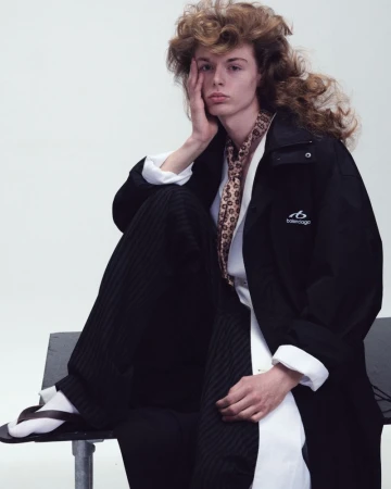 Model Nik Dermendzhiev wearing a black Balenciaga coat, patterned scarf, and white shirt seated in front of a white studio backdrop.