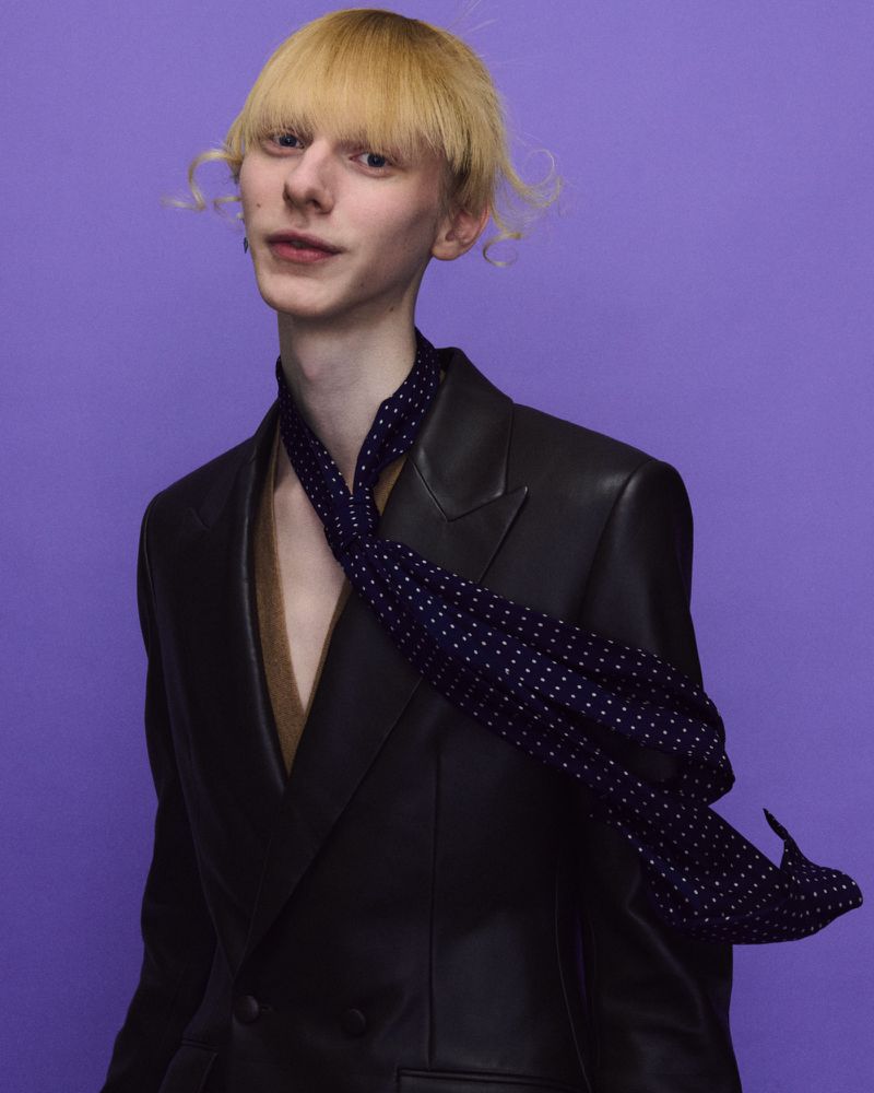 A blonde model wearing a black leather blazer and polka dot skinny scarf posing against a solid purple backdrop.