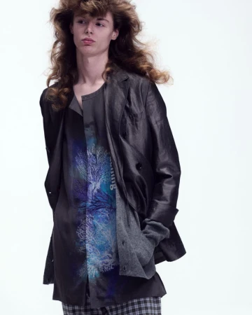 Model Nik Dermendzhiev with voluminous wavy hair wearing a black layered jacket and graphic print shirt against a white background.