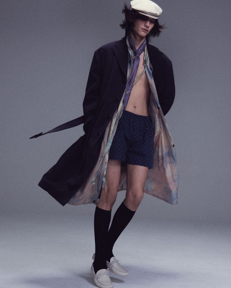 Model Nik Dermendzhiev wearing an open black overcoat, patterned shorts, flat cap, and white loafers against a studio backdrop.