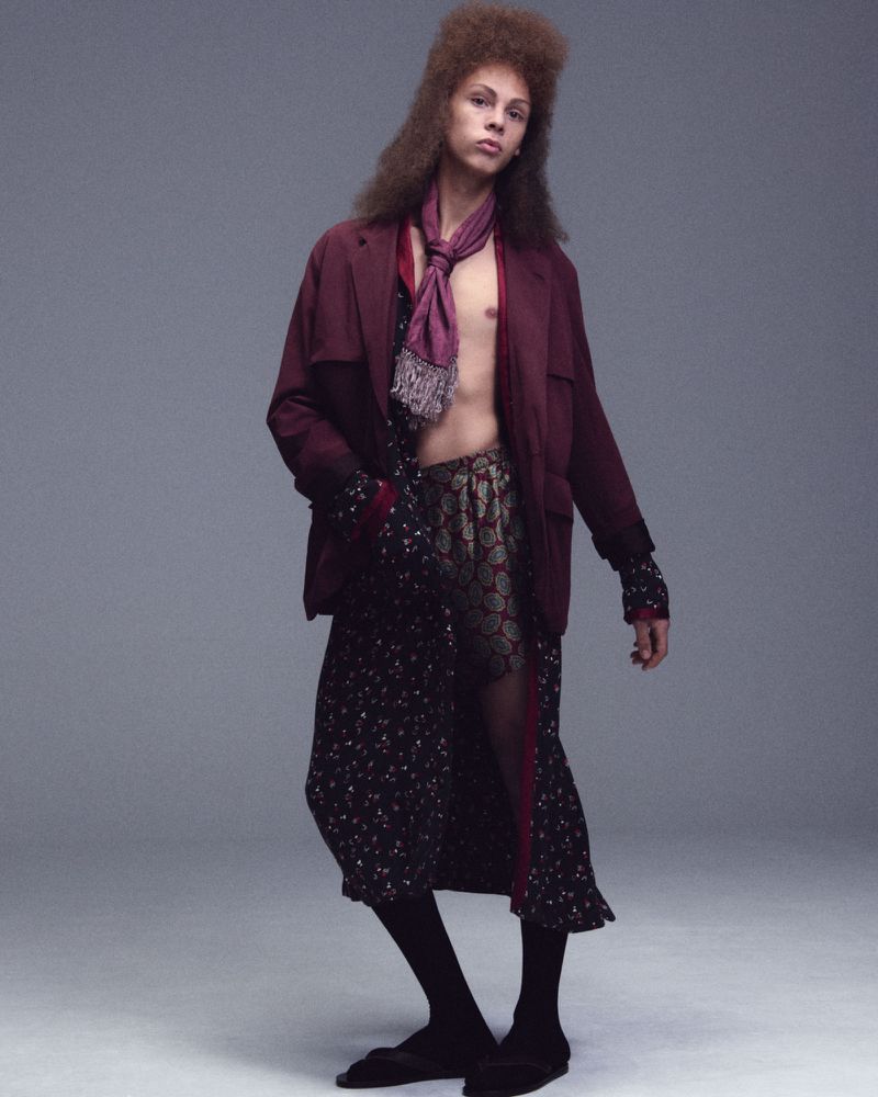 Fashionable adult male in a burgundy jacket, patterned dress, and socks, posing in a studio environment with neutral grey background.