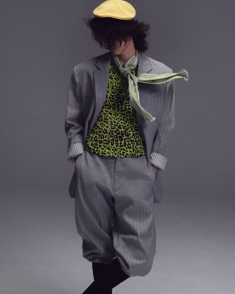 Model with dark hair wearing a grey suit, green leopard print top, yellow hat, and striped scarf against a plain grey backdrop.