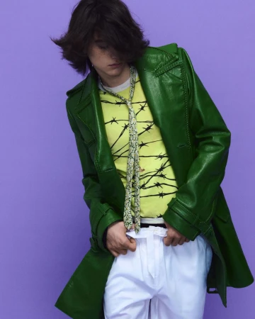 Model Nik Dermendzhiev wearing a green leather trench coat, yellow graphic tee, and white pants against a lavender background.