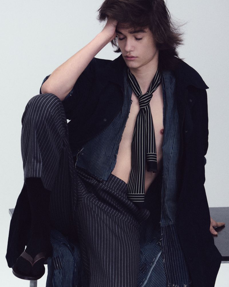 Male model seated with hand on head wearing striped shirt, necktie, and dark coat against a plain background
