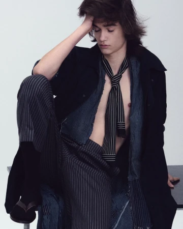 Male model seated with hand on head wearing striped shirt, necktie, and dark coat against a plain background