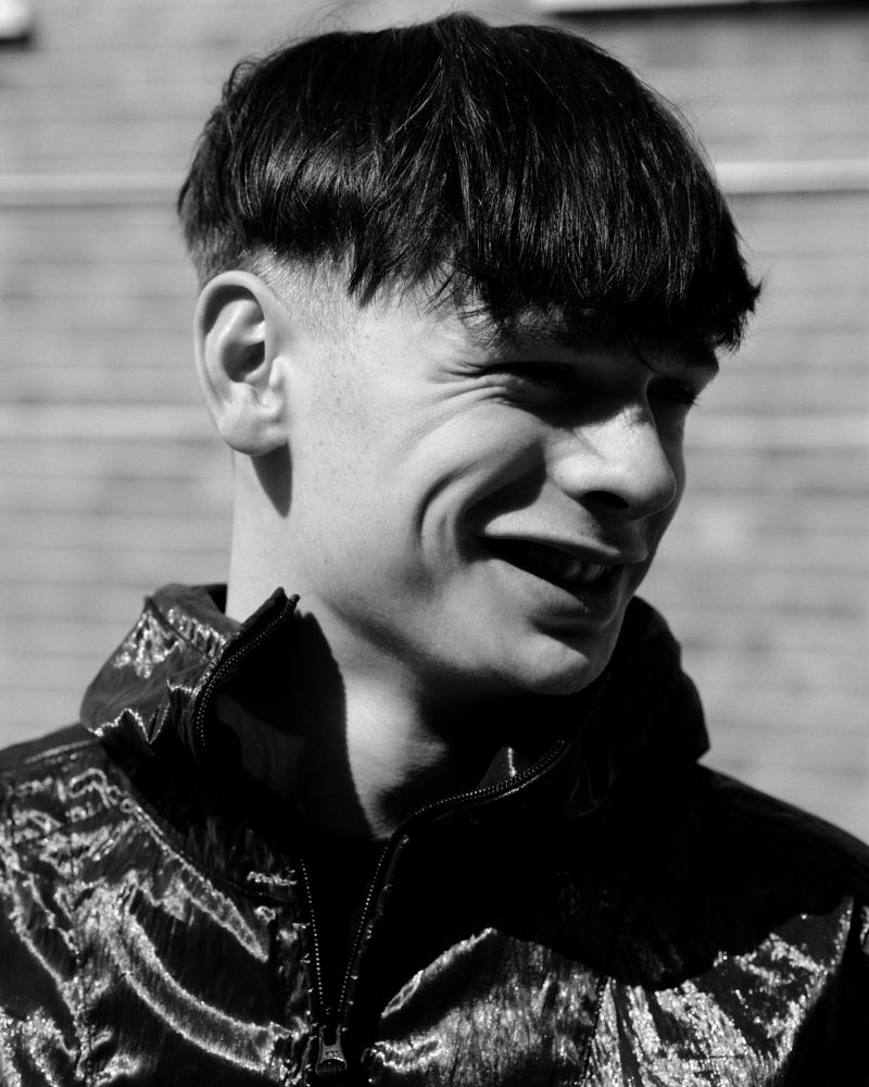 Portrait of a young man with dark bowl cut hair wearing a shiny zipped jacket outdoors in black and white