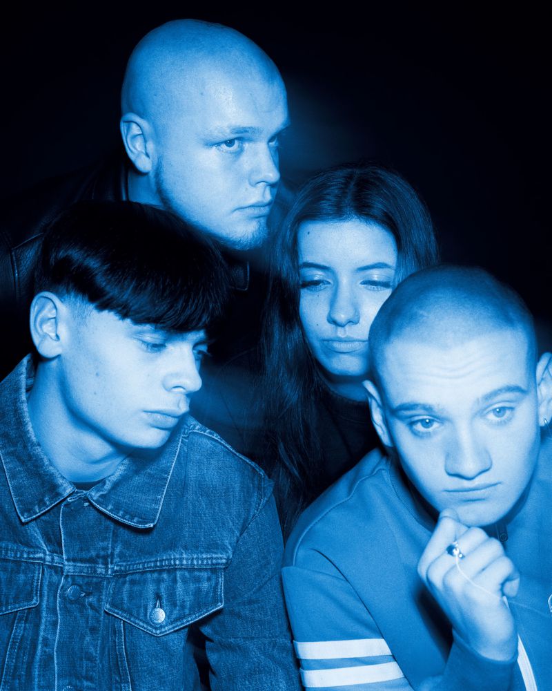 Four young people with distinct haircuts illuminated in deep blue light against a black backdrop in a group portrait.