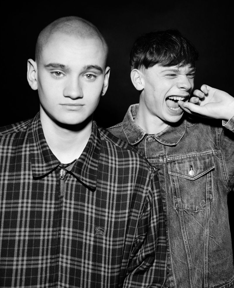 Black and white portrait of two young men, one in a plaid zip jacket and another laughing in a denim jacket