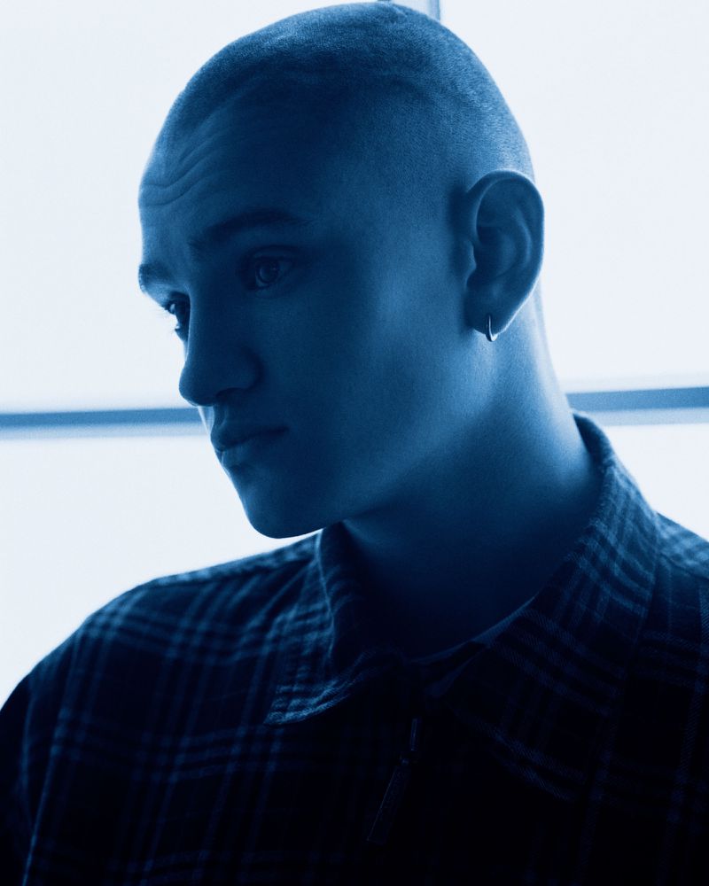 Close-up portrait of a young man with a buzz cut wearing a plaid shirt in cool blue monochrome lighting.