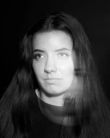 Monochrome portrait of a young woman with long dark hair wearing a dark crewneck sweater against a dark background.