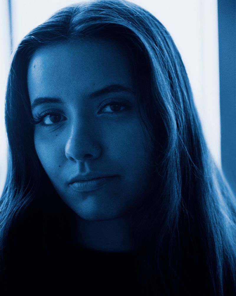 Blue-toned close-up portrait of a young woman with long dark hair looking directly at the camera.