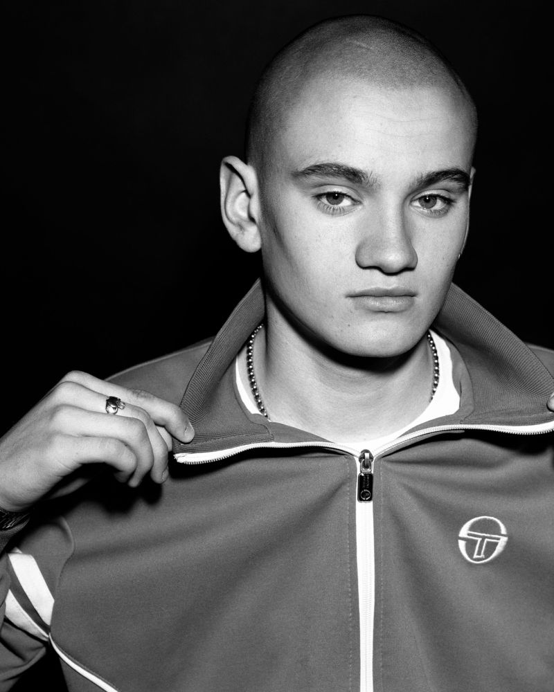 Black and white portrait of a young man with a shaved head wearing a zip-up track jacket and chain necklace against a dark background.