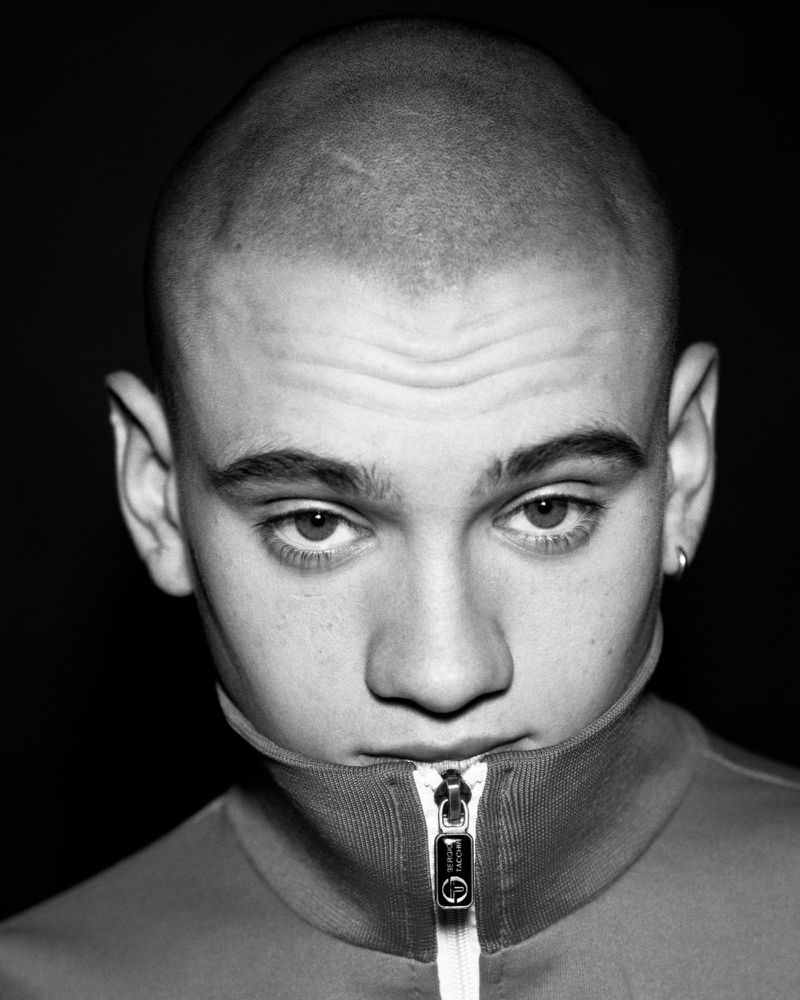 Close up portrait of a young man with a shaved head wearing a high collared zip up jacket against a dark background