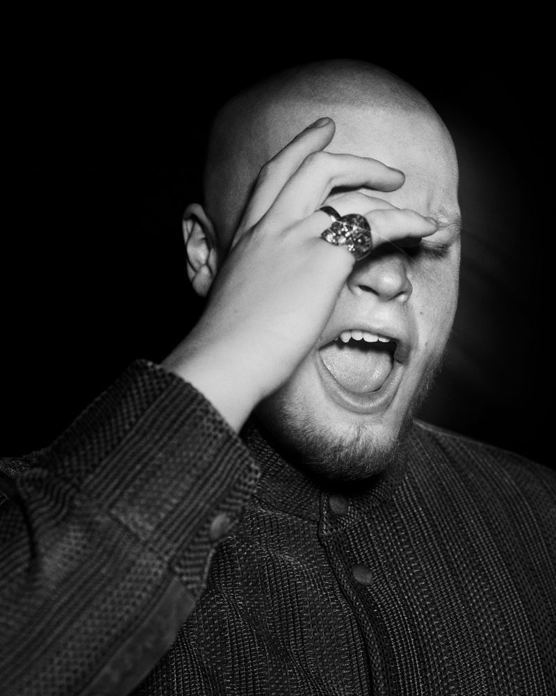 Black and white portrait of a bald man with a beard wearing a buttoned shirt, with his hand near his face and mouth open in photo studio.