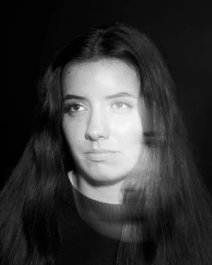 Monochrome portrait of a young woman with long dark hair wearing a dark crewneck sweater against a dark background.