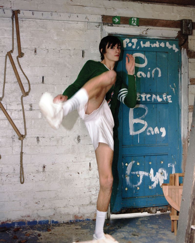 A person with dark hair in a green sweater and white shorts kicks one leg up in an indoor location in front of a blue door with graffiti.