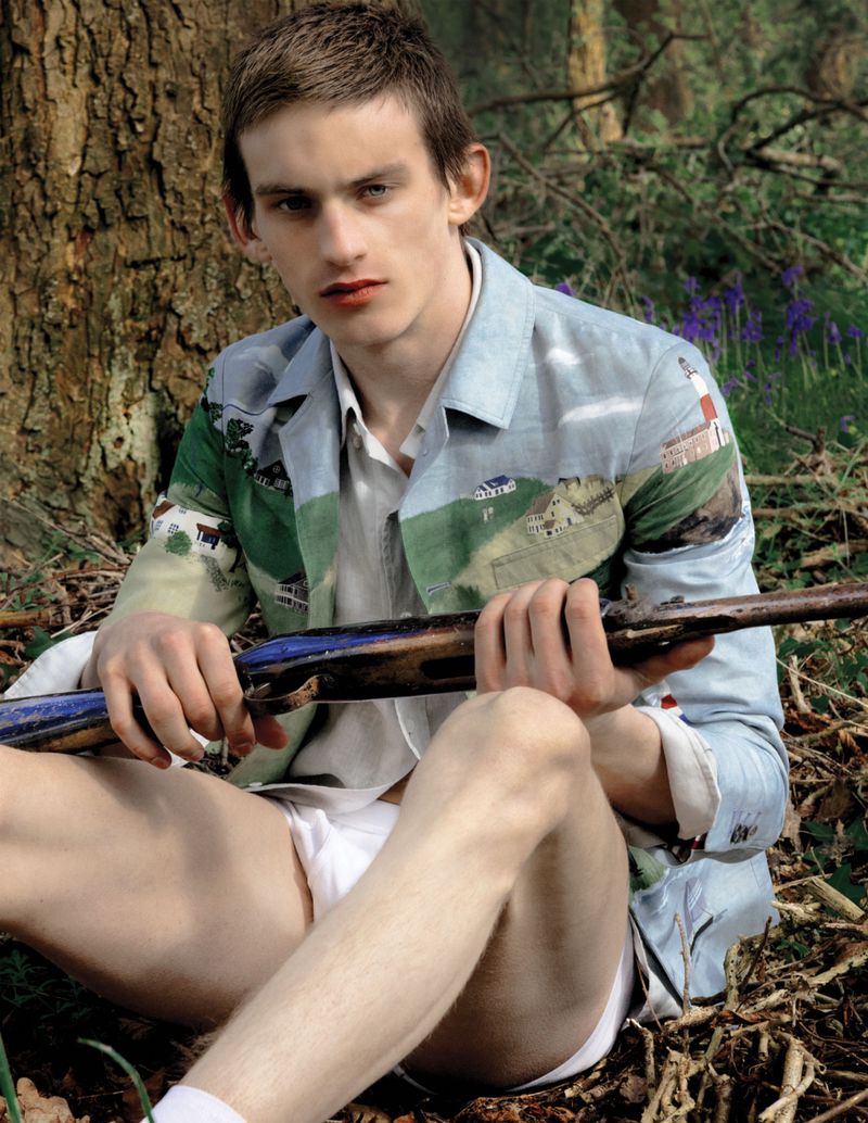 Model Joe Bottomer sits against a tree in a woodland setting wearing a landscape-printed blazer and holding a vintage rifle.