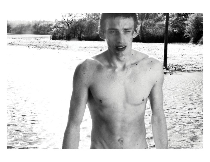 Black and white portrait of a shirtless young man standing in a body of water outdoors with trees in the background.
