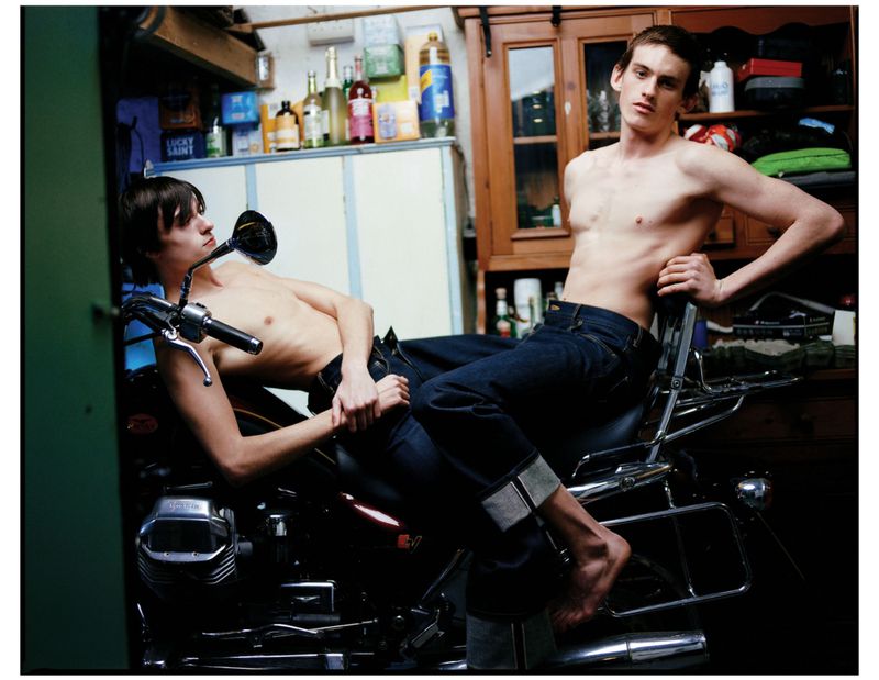 Two shirtless young men in dark denim jeans lounging on a vintage motorcycle inside a cluttered room with wooden cabinets.